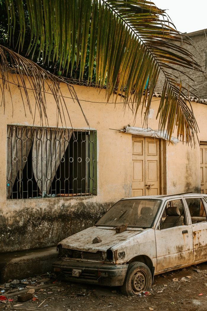 the-story A rustic scene of a damaged car parked by an old house with barred windows and palm fronds.